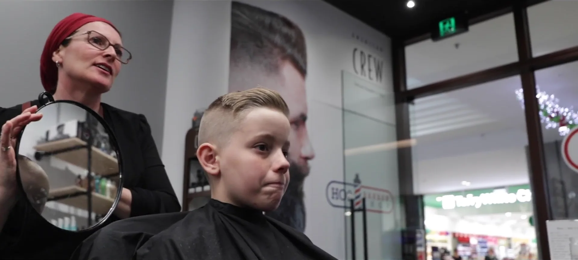 A barber holds up the hand mirror so a young customer can see the back of his fade